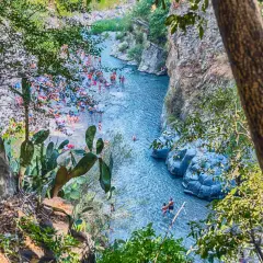 Gente si gode il sole e l'acqua cristallina nella Valle dell'Alcantara, tra rocce vulcaniche e vegetazione rigogliosa.