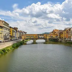 Il Ponte Vecchio, con le sue botteghe colorate, si specchia sull'Arno. Un canoista solitario solca le acque sotto il cielo azzurro e le nuvole bianche.