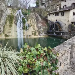 Antico mulino ad acqua accanto a cascate e grotte in Veneto, con un laghetto verde e vegetazione rigogliosa.