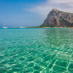 Acque cristalline e turchesi bagnano la spiaggia di San Vito Lo Capo, con il Monte Monaco imponente sullo sfondo. Velieri solcano il mare azzurro.