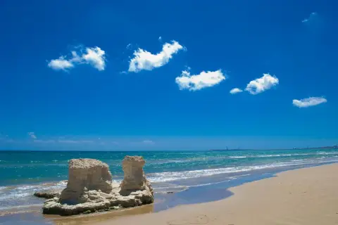 Spiagge attrezzate Sicilia: rocce scolpite dal mare e sabbia dorata sotto un cielo azzurro con nuvole bianche.