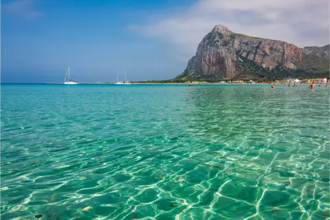 Acque cristalline e turchesi bagnano la spiaggia di San Vito Lo Capo, con il Monte Monaco imponente sullo sfondo. Velieri solcano il mare azzurro.