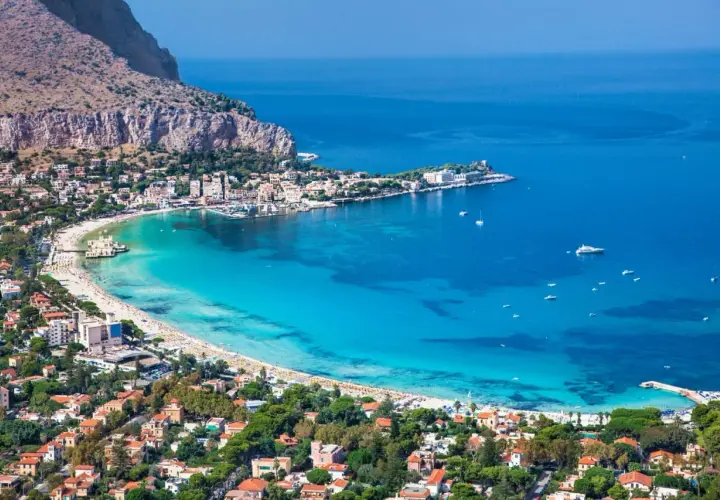 Vista aerea della spiaggia di Palermo, con acque turchesi, case colorate e montagne sullo sfondo.