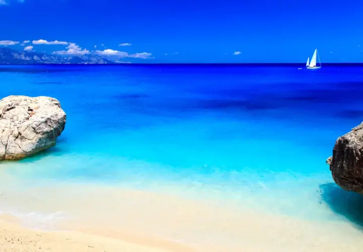 Spiaggia dorata bagnata da un mare cristallino dalle sfumature turchesi e blu intenso. In lontananza, una barca a vela solca l'orizzonte.