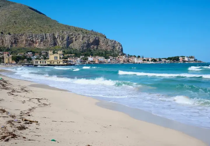 Spiaggia dorata e mare turchese in Sicilia, con un borgo colorato incastonato tra la montagna e l'acqua cristallina.