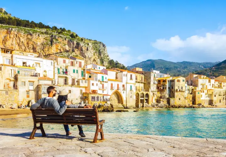 Coppia seduta su una panchina, ammira il mare e le case colorate di Cefalù, un luogo perfetto per vacanze in Sicilia sul mare.