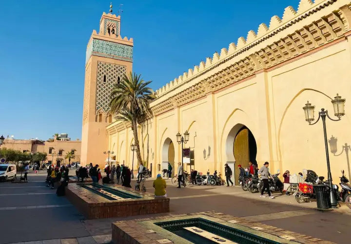 La moschea Koutoubia domina la piazza, con la sua torre decorata e le arcate del souk di Marrakech. Gente e motorini animano la scena.