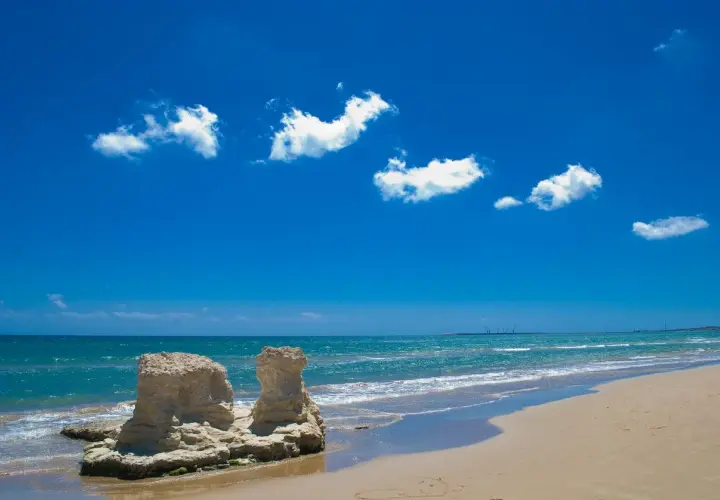 Spiagge attrezzate Sicilia: rocce scolpite dal mare e sabbia dorata sotto un cielo azzurro con nuvole bianche.