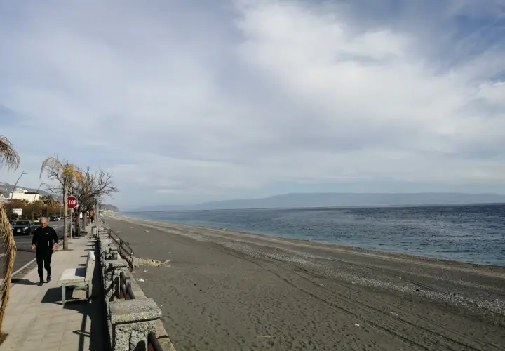 Un uomo corre sul lungomare di Roccafiorita, la spiaggia scura si estende verso l'orizzonte sotto un cielo nuvoloso.