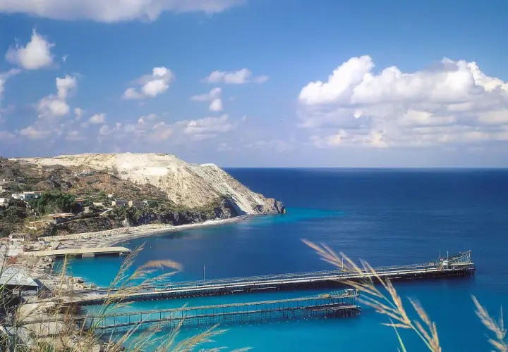 Panoramica delle isole Lipari con un porto e moli che si estendono nel mare blu intenso sotto un cielo azzurro con nuvole bianche.