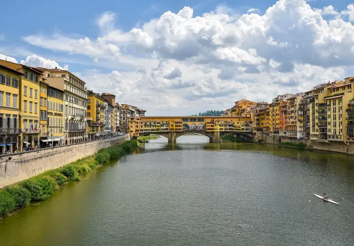 Il Ponte Vecchio, con le sue botteghe colorate, si specchia sull'Arno. Un canoista solitario solca le acque sotto il cielo azzurro e le nuvole bianche.