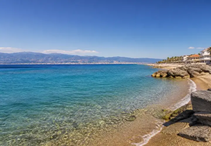Spiaggia di Reggio Calabria con mare cristallino, palme e case bianche. All'orizzonte, montagne azzurre sotto un cielo limpido.
