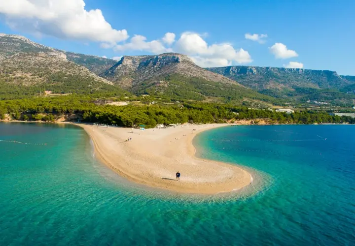 Una spiaggia a forma di lingua di sabbia dorata si estende nel mare turchese della costa croata, con montagne verdi sullo sfondo.