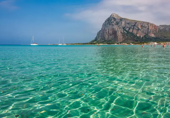 Acque cristalline e turchesi bagnano la spiaggia di San Vito Lo Capo, con il Monte Monaco imponente sullo sfondo. Velieri solcano il mare azzurro.