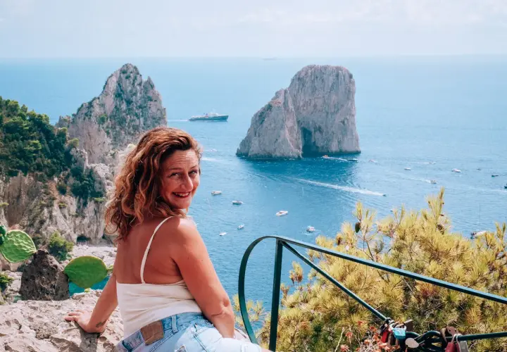 Donna sorridente ammira i Faraglioni, una delle cose da vedere a Capri, con il mare azzurro e barche.