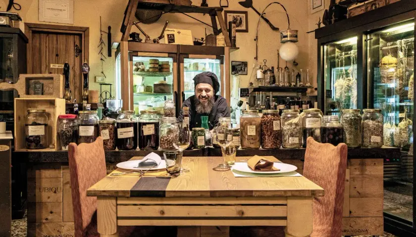 Un cuoco sorridente in un locale con salumi appesi e barattoli di conserve. Un'atmosfera unica per ristoranti particolari a Brescia e provincia.