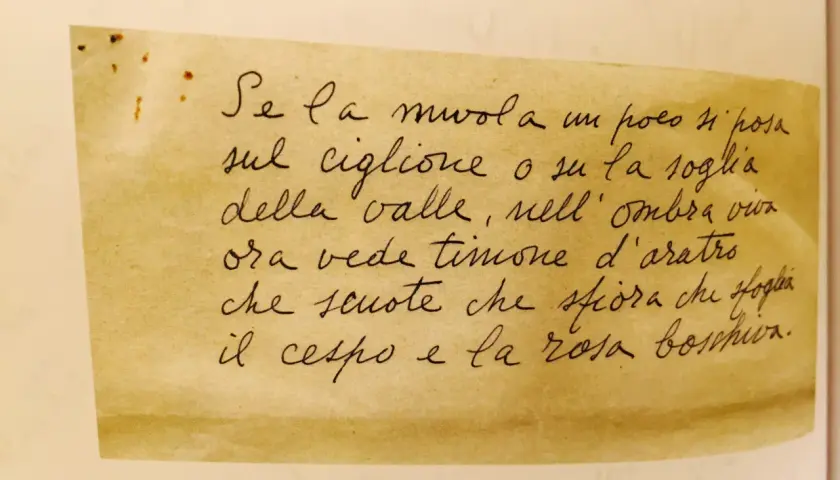 Scrittura a mano su carta ingiallita: "Se la nuvola un poco si posa sul ciglione o su la soglia della valle, nell'ombra viva ora vede timone d'aratro che scuote che sfiora che sfoglia il cespo e la rosa boschiva." Un piccolo lucio.