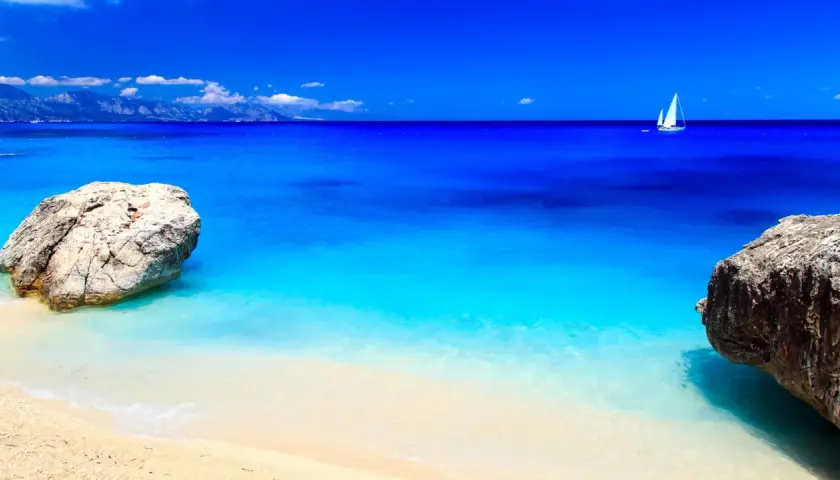 Spiaggia dorata bagnata da un mare cristallino dalle sfumature turchesi e blu intenso. In lontananza, una barca a vela solca l'orizzonte.