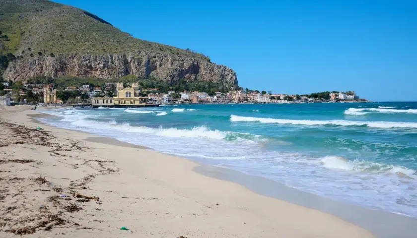 Spiaggia dorata e mare turchese in Sicilia, con un borgo colorato incastonato tra la montagna e l'acqua cristallina.
