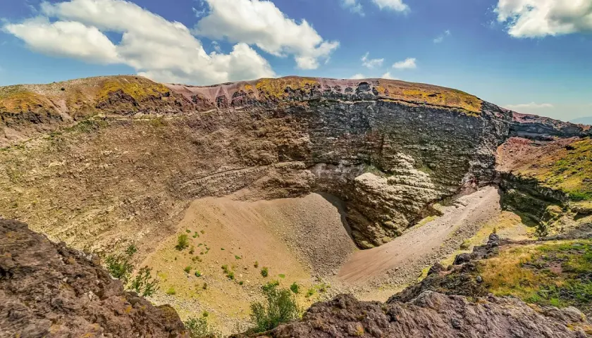 Il cratere del Vesuvio, con le sue pareti stratificate e il cono di detriti al centro, sotto un cielo azzurro con nuvole bianche.