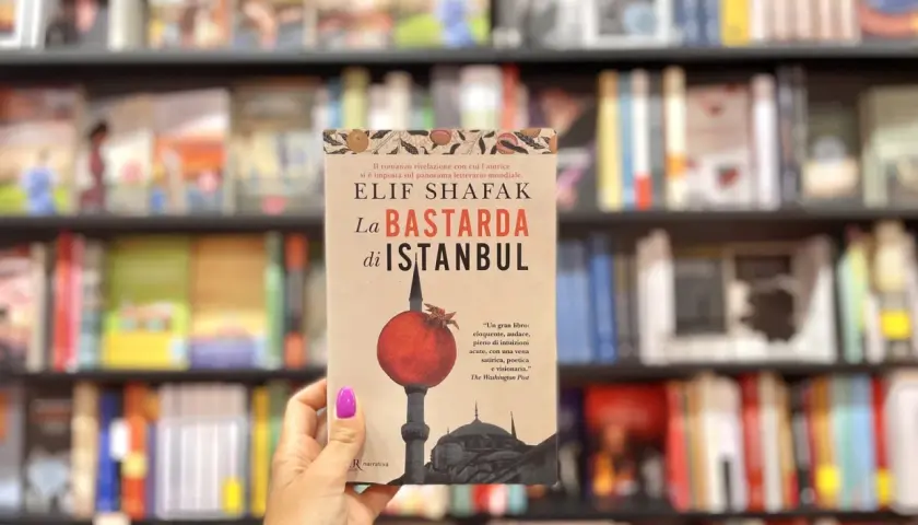 Mano tiene "La Bastarda di Istanbul", un libro che esplora le storie di Istanbul, tra scaffali pieni di libri.