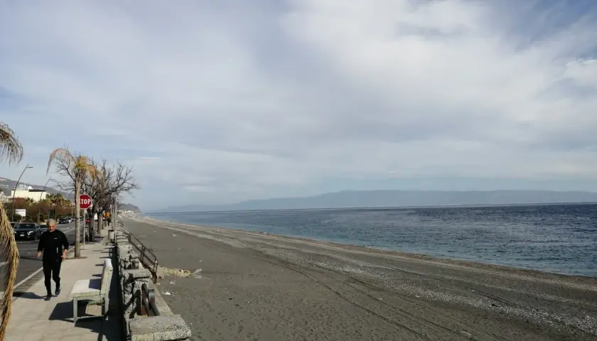 Un uomo corre sul lungomare di Roccafiorita, la spiaggia scura si estende verso l'orizzonte sotto un cielo nuvoloso.