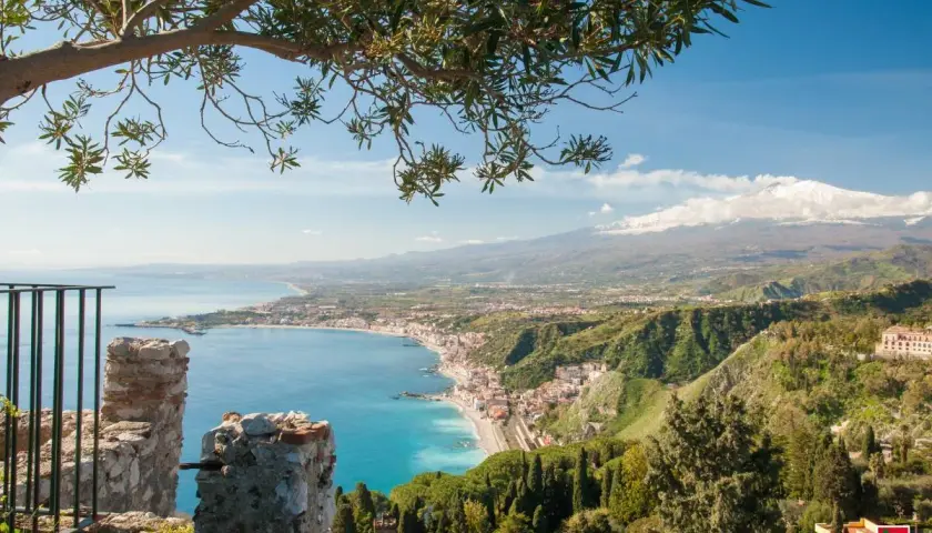 Vista panoramica di Naxos, Sicilia, con il mare azzurro, la costa e l'Etna innevato sullo sfondo, incorniciata da un ramo d'ulivo.