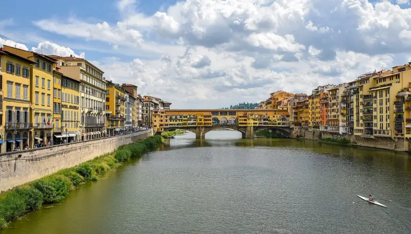 Il Ponte Vecchio, con le sue botteghe colorate, si specchia sull'Arno. Un canoista solitario solca le acque sotto il cielo azzurro e le nuvole bianche.