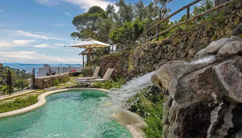 Piscina con cascata che ricorda le terme naturali di Ischia, circondata da vegetazione e con vista sul mare.