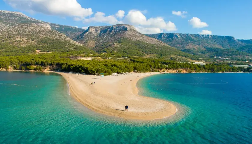 Una spiaggia a forma di lingua di sabbia dorata si estende nel mare turchese della costa croata, con montagne verdi sullo sfondo.
