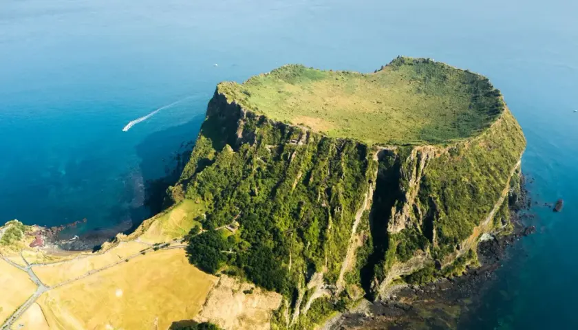 Un cratere vulcanico verdeggiante, tra le meraviglie naturali del mondo, si erge dal mare blu. Una barca solca le acque.