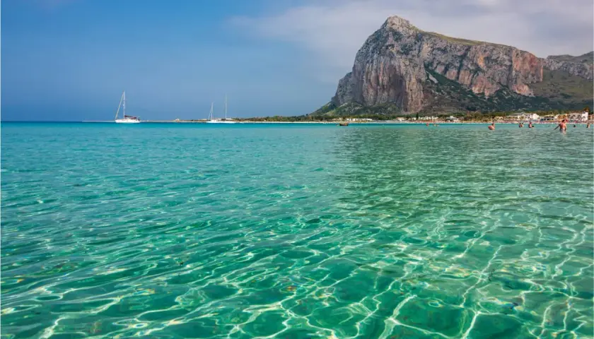 Acque cristalline e turchesi bagnano la spiaggia di San Vito Lo Capo, con il Monte Monaco imponente sullo sfondo. Velieri solcano il mare azzurro.