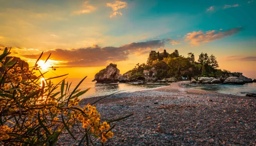 Tramonto dorato sulle spiagge di Giardini Naxos Taormina, con rocce e vegetazione lussureggiante.
