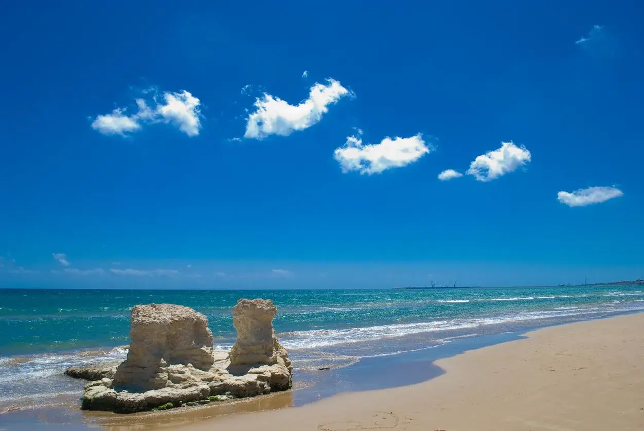 Spiagge attrezzate Sicilia: rocce scolpite dal mare e sabbia dorata sotto un cielo azzurro con nuvole bianche.