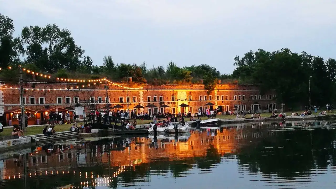 Serata estiva a Marghera: cosa vedere? Un forte storico illuminato da lucine, con gente seduta ai tavolini e barche ormeggiate.