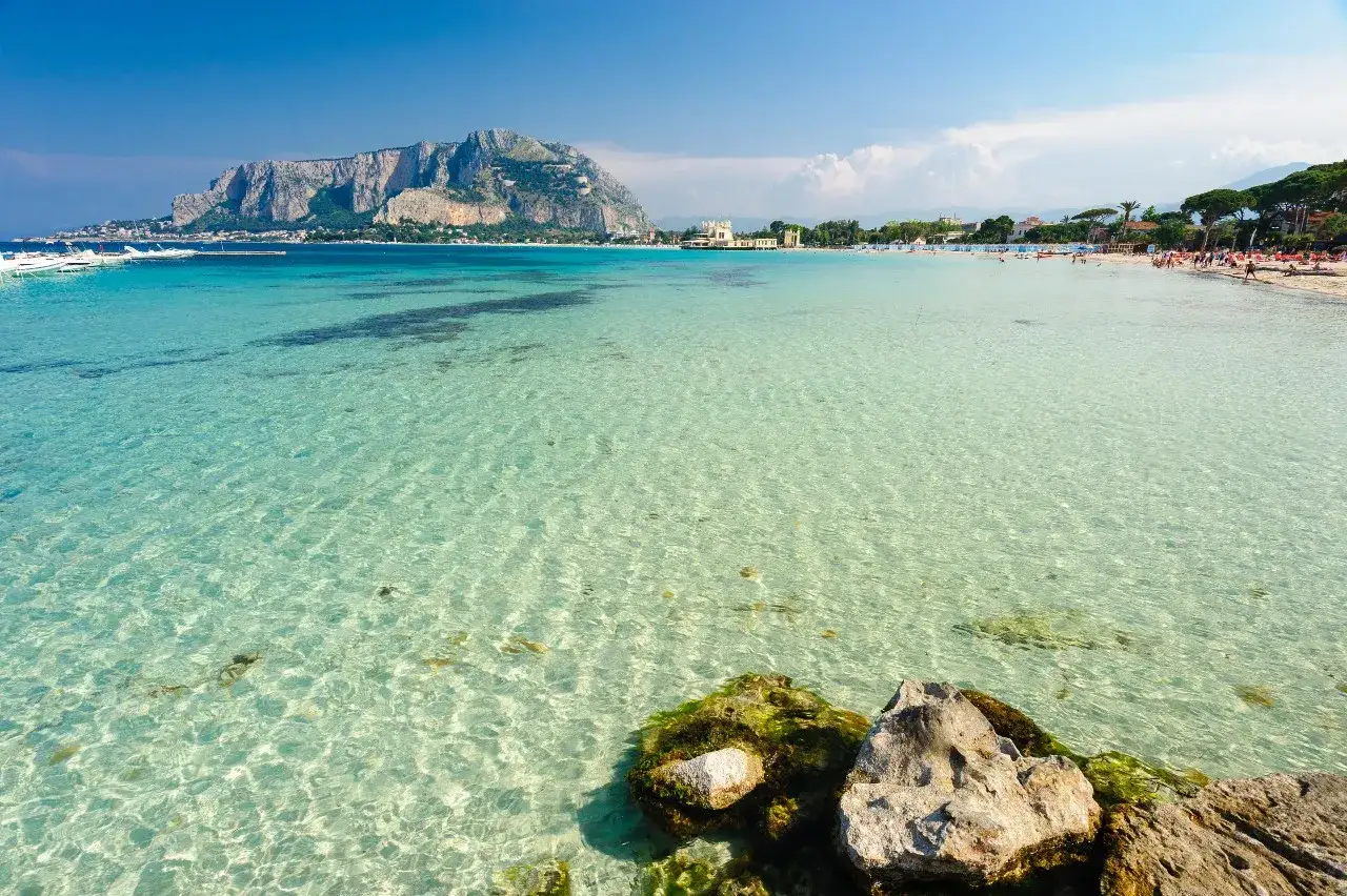 Acque cristalline e un promontorio maestoso fanno da cornice a questo splendido mare a Palermo, con barche ormeggiate e bagnanti che godono del sole.