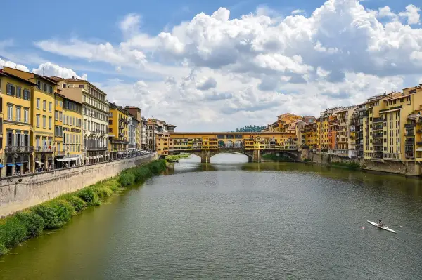 Il Ponte Vecchio, con le sue botteghe colorate, si specchia sull'Arno. Un canoista solitario solca le acque sotto il cielo azzurro e le nuvole bianche.